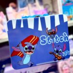 Valisette métallique de rangement bleu vif illustrée avec le personnage Disney Stitch dans trois poses : portant une planche de surf rouge, jouant de la guitare avec des notes de musique, et portant des lunettes de soleil roses avec une fleur. La boîte possède une poignée bleue et un fermoir en métal argenté.