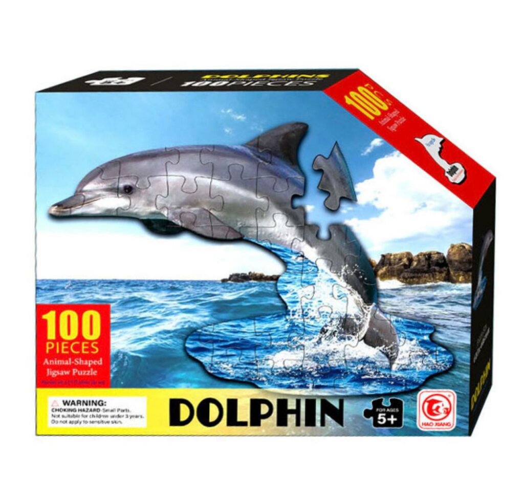 Boîte de puzzle rectangulaire montrant un dauphin gris en pièces de puzzle sautant au-dessus d'une mer bleue. Le texte indique "100 PIECES", "Animal-Shaped Jigsaw Puzzle", "DOLPHIN" et "FOR AGES 5+"
