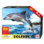 Boîte de puzzle rectangulaire montrant un dauphin gris en pièces de puzzle sautant au-dessus d'une mer bleue. Le texte indique "100 PIECES", "Animal-Shaped Jigsaw Puzzle", "DOLPHIN" et "FOR AGES 5+"
