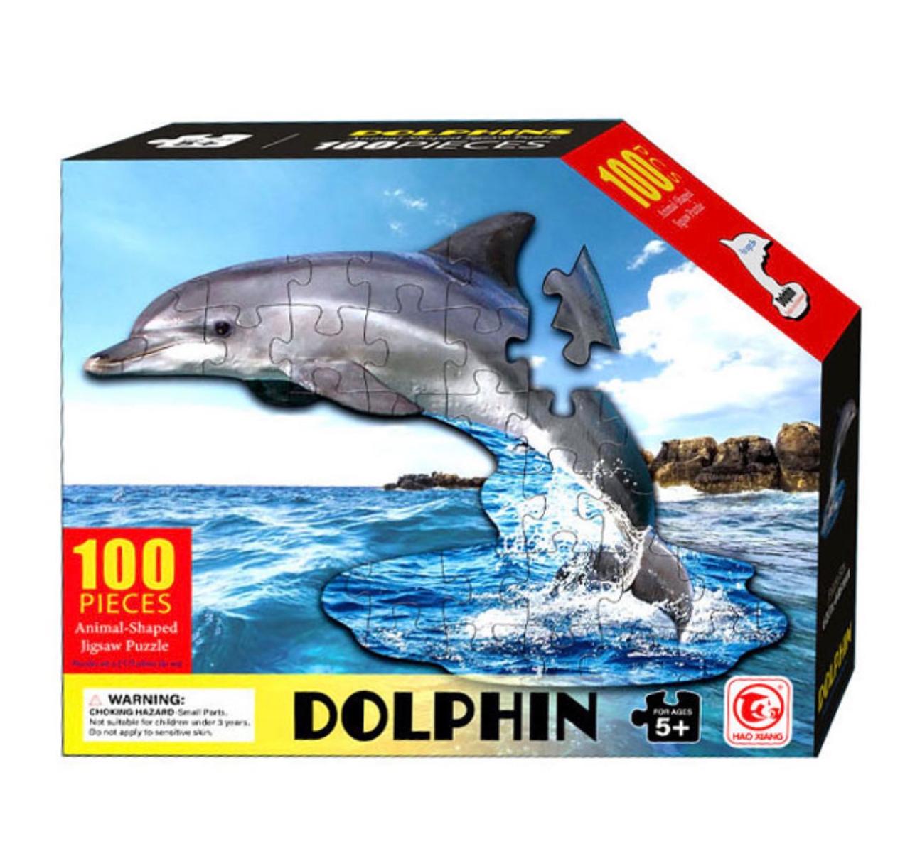 Boîte de puzzle rectangulaire montrant un dauphin gris en pièces de puzzle sautant au-dessus d'une mer bleue. Le texte indique "100 PIECES", "Animal-Shaped Jigsaw Puzzle", "DOLPHIN" et "FOR AGES 5+"