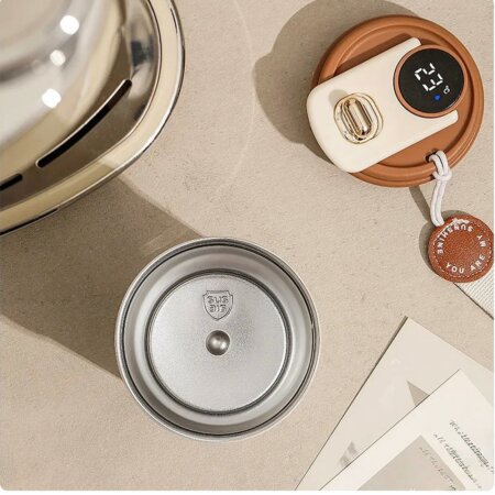 Vue de dessus d'un mug isotherme ouvert posé sur une surface texturée beige. On aperçoit l'intérieur de la tasse en acier inoxydable argenté marqué du sceau "SUS 316". À côté, le couvercle marron et crème affiche "23°C" sur un écran numérique LED