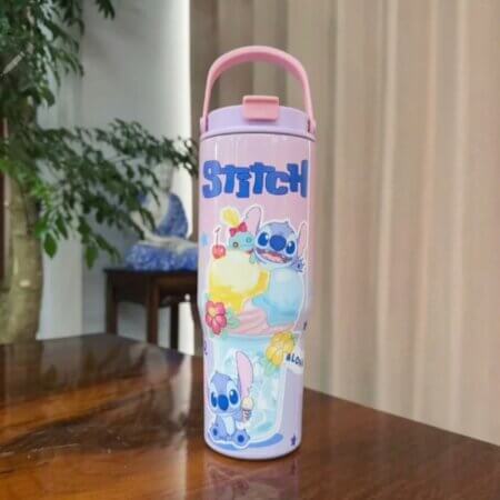 Mug isotherme de grande capacité au design dégradé rose et mauve, illustré avec le personnage de Disney Stitch et des motifs tropicaux Aloha. La tasse en acier inoxydable possède un couvercle rose avec une poignée de transport intégrée, posée sur une table en bois brun. »
