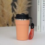 mug isotherme de voyage au design moderne et épuré. Le gobelet, de couleur pêche pastel, est doté d'une finition mate et d'une structure en acier inoxydable visible au niveau du bord supérieur.