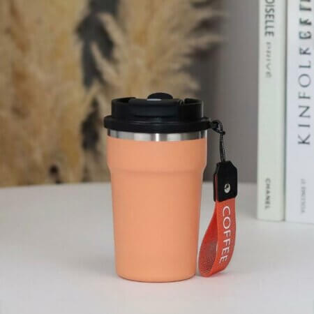 mug isotherme de voyage au design moderne et épuré. Le gobelet, de couleur pêche pastel, est doté d'une finition mate et d'une structure en acier inoxydable visible au niveau du bord supérieur.