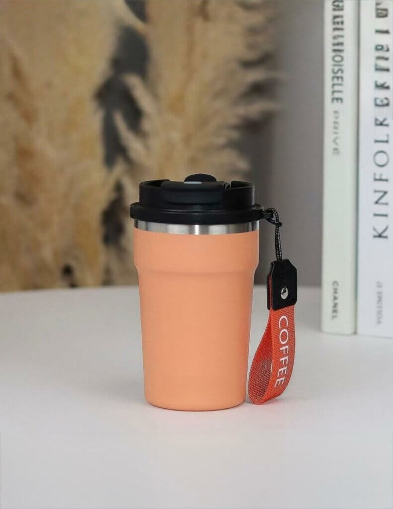 mug isotherme de voyage au design moderne et épuré. Le gobelet, de couleur pêche pastel, est doté d'une finition mate et d'une structure en acier inoxydable visible au niveau du bord supérieur.
