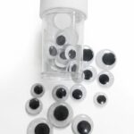 des yeux mobiles classiques (souvent appelés "googly eyes" en anglais), présentés dans un petit flacon transparent avec un bouchon blanc. Contrairement à l'assortiment multicolore vu précédemment, ceux-ci sont le modèle standard : une base blanche, une capsule transparente et une pupille noire mobile à l'intérieur.