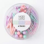 Un organisateur de bureau circulaire transparent contenant quatre compartiments distincts remplis de fournitures pastel : des punaises à tête plate, des trombones en forme de goutte, des trombones standards et des punaises sphériques