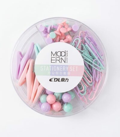 Un organisateur de bureau circulaire transparent contenant quatre compartiments distincts remplis de fournitures pastel : des punaises à tête plate, des trombones en forme de goutte, des trombones standards et des punaises sphériques