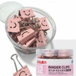 Boîte ronde transparente remplie de pinces à dessin rose pastel avec un motif de visage souriant (smiley) découpé, accompagnées d'une pince posée à côté sur un fond blanc.