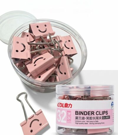 Boîte ronde transparente remplie de pinces à dessin rose pastel avec un motif de visage souriant (smiley) découpé, accompagnées d'une pince posée à côté sur un fond blanc.