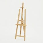 Un chevalet de peinture en bois clair de style trépied, vide, sur un fond blanc uni, montrant sa structure réglable.