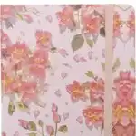 Un carnet de notes fermé avec une couverture rigide rose clair ornée d'un motif floral délicat représentant des fleurs dans les tons rose vif, saumon et jaune avec de petites feuilles vertes. Une bande élastique verticale de couleur rose pâle est fixée sur le côté droit pour maintenir le carnet bien fermé.