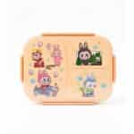 Une boîte à déjeuner (lunch box) pour enfant de couleur beige clair, encore sous emballage plastique transparent. Le couvercle est divisé visuellement en plusieurs zones décorées d'illustrations de style bande dessinée ou manga
