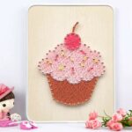 Un tableau en art filaire représentant un cupcake coloré sur une planche en bois clair. La base du gâteau est tissée avec des fils marrons, le glaçage est en fils rose clair parsemé de petites touches jaunes pour imiter des vermicelles, et le sommet est surmonté d'une cerise en fils rose foncé.