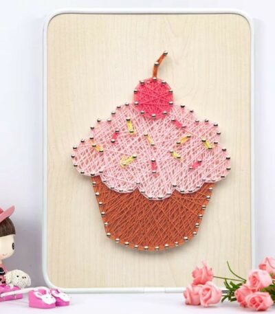 Un tableau en art filaire représentant un cupcake coloré sur une planche en bois clair. La base du gâteau est tissée avec des fils marrons, le glaçage est en fils rose clair parsemé de petites touches jaunes pour imiter des vermicelles, et le sommet est surmonté d'une cerise en fils rose foncé.