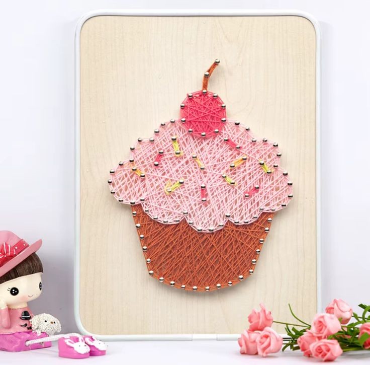 Un tableau en art filaire représentant un cupcake coloré sur une planche en bois clair. La base du gâteau est tissée avec des fils marrons, le glaçage est en fils rose clair parsemé de petites touches jaunes pour imiter des vermicelles, et le sommet est surmonté d'une cerise en fils rose foncé.