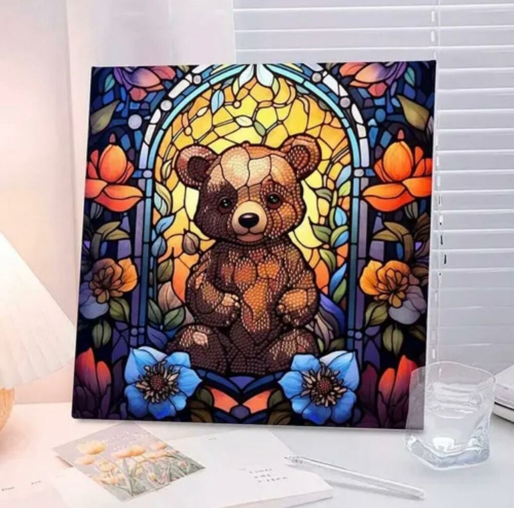 Un tableau carré de broderie diamant, sans cadre visible, est exposé sur un bureau près d'une fenêtre à stores blancs. L'œuvre représente un ourson brun assis au centre, stylisé dans un esprit vitrail avec des textures perlées. Il se détache sur un fond jaune doré lumineux, encadré par des arches bleues et entouré de grandes fleurs colorées (bleues au premier plan, orange et violettes sur les côtés). Un verre d'eau et des carnets ouverts entourent le tableau.