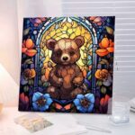 Un tableau carré de broderie diamant, sans cadre visible, est exposé sur un bureau près d'une fenêtre à stores blancs. L'œuvre représente un ourson brun assis au centre, stylisé dans un esprit vitrail avec des textures perlées. Il se détache sur un fond jaune doré lumineux, encadré par des arches bleues et entouré de grandes fleurs colorées (bleues au premier plan, orange et violettes sur les côtés). Un verre d'eau et des carnets ouverts entourent le tableau.