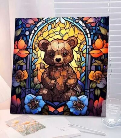Un tableau carré de broderie diamant, sans cadre visible, est exposé sur un bureau près d'une fenêtre à stores blancs. L'œuvre représente un ourson brun assis au centre, stylisé dans un esprit vitrail avec des textures perlées. Il se détache sur un fond jaune doré lumineux, encadré par des arches bleues et entouré de grandes fleurs colorées (bleues au premier plan, orange et violettes sur les côtés). Un verre d'eau et des carnets ouverts entourent le tableau.