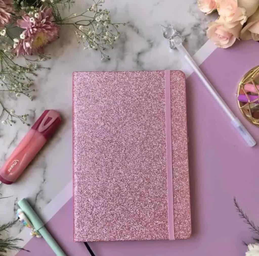 Un carnet de notes format A5 fermé, doté d'une couverture rigide entièrement recouverte de paillettes rose vif scintillantes. Une bande élastique verticale rose assortie est fixée sur le côté droit pour maintenir le carnet fermé. L'ensemble est photographié de face sur un fond blanc uni.
