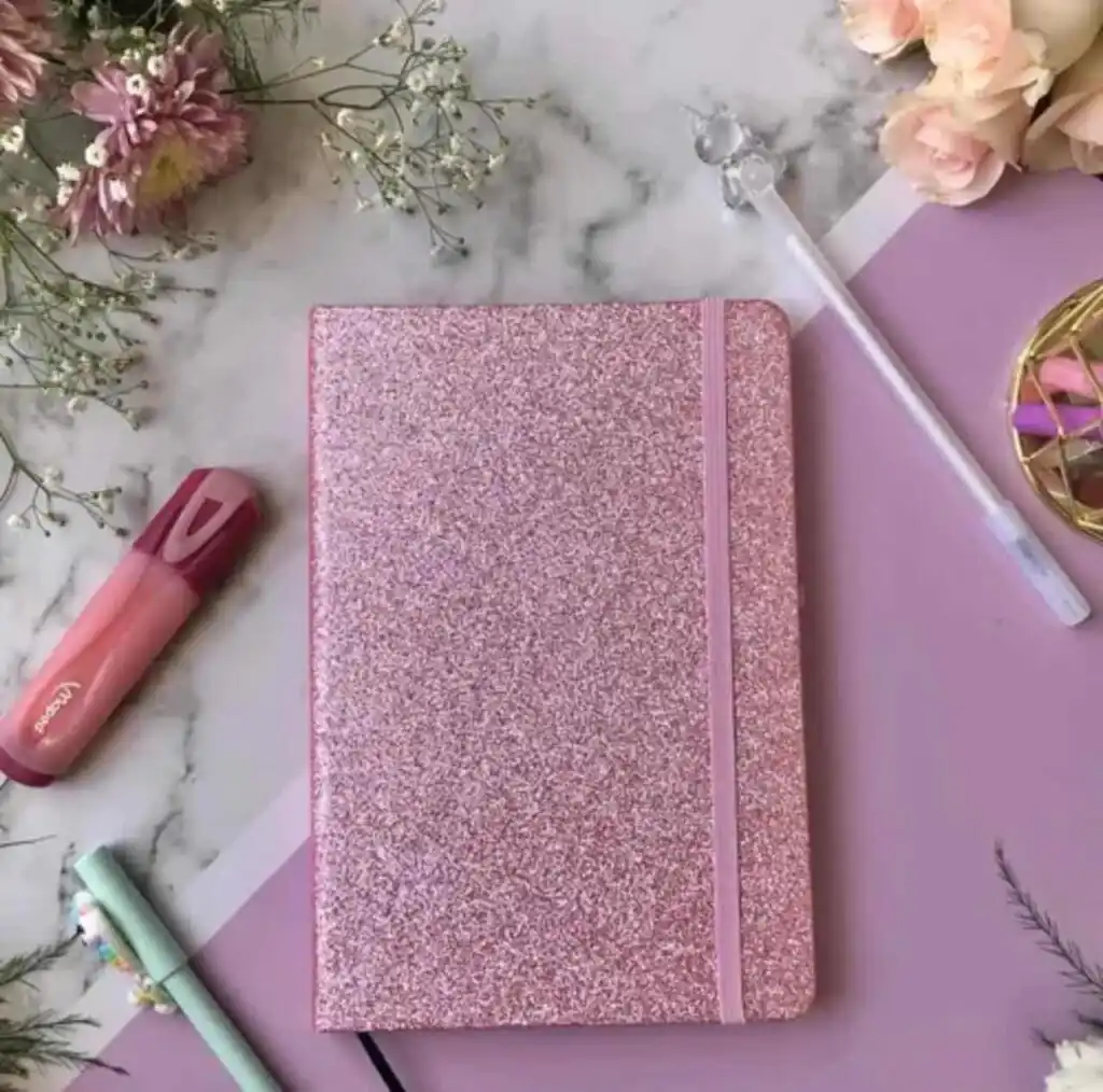 Un carnet de notes format A5 fermé, doté d'une couverture rigide entièrement recouverte de paillettes rose vif scintillantes. Une bande élastique verticale rose assortie est fixée sur le côté droit pour maintenir le carnet fermé. L'ensemble est photographié de face sur un fond blanc uni.