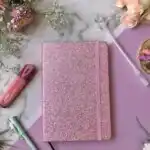 Un carnet de notes format A5 fermé, doté d'une couverture rigide entièrement recouverte de paillettes rose vif scintillantes. Une bande élastique verticale rose assortie est fixée sur le côté droit pour maintenir le carnet fermé. L'ensemble est photographié de face sur un fond blanc uni.