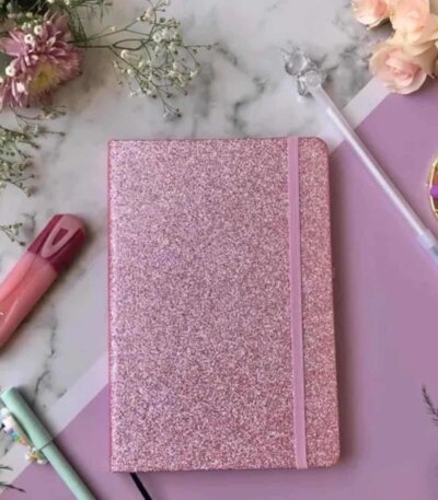 Un carnet de notes format A5 fermé, doté d'une couverture rigide entièrement recouverte de paillettes rose vif scintillantes. Une bande élastique verticale rose assortie est fixée sur le côté droit pour maintenir le carnet fermé. L'ensemble est photographié de face sur un fond blanc uni.