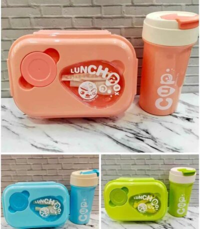 Une boîte à lunch rectangulaire aux bords arrondis avec un couvercle transparent au centre, laissant voir des couverts (fourchette et cuillère) et une petite illustration de personnage. Le mot "LUNCH Box" est inscrit en arc de cercle sur le couvercle.