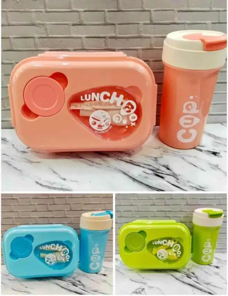Une boîte à lunch rectangulaire aux bords arrondis avec un couvercle transparent au centre, laissant voir des couverts (fourchette et cuillère) et une petite illustration de personnage. Le mot "LUNCH Box" est inscrit en arc de cercle sur le couvercle.