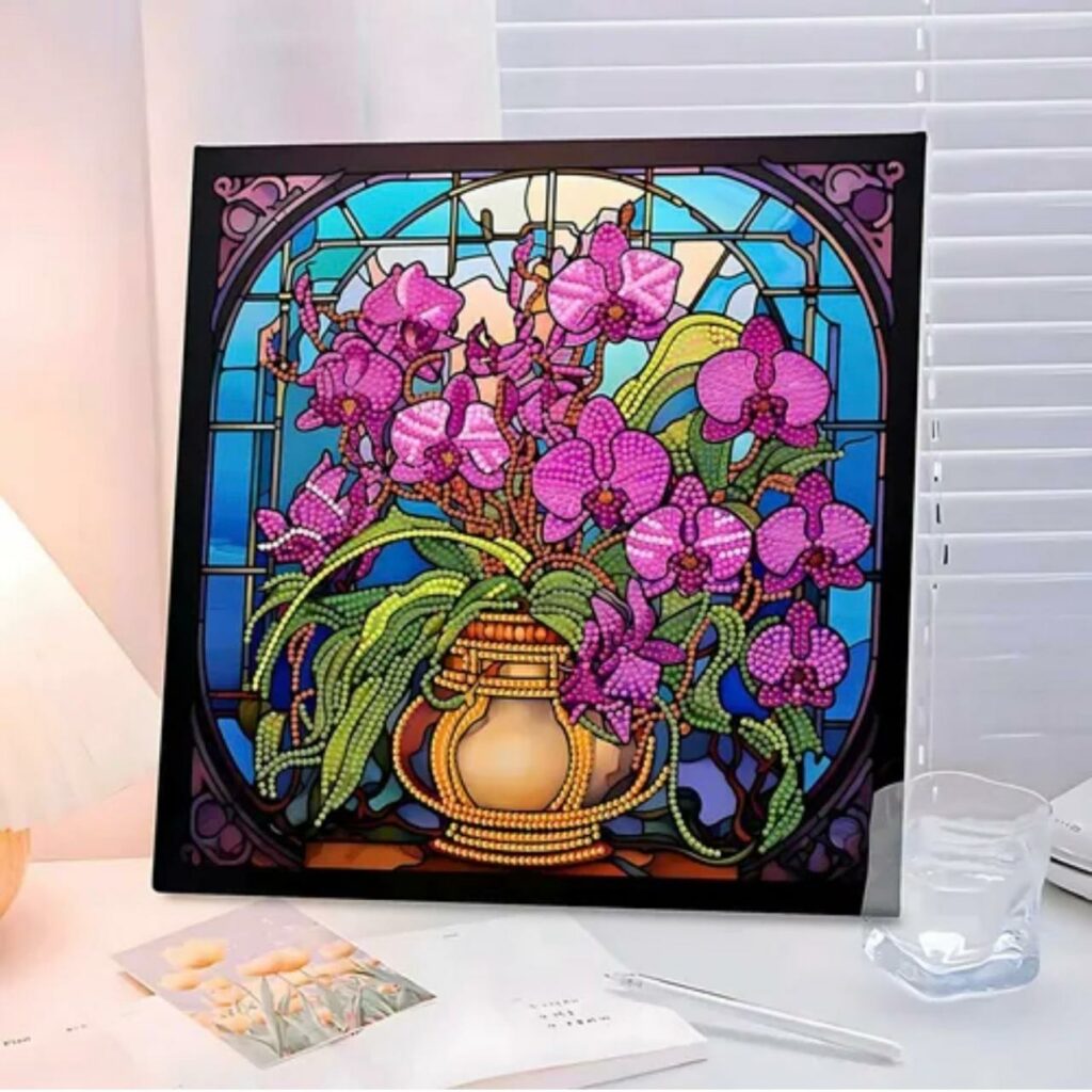 Un tableau de broderie diamant carré, inséré dans un cadre noir, est exposé sur un bureau éclairé. L'œuvre représente un vase doré contenant un grand bouquet d'orchidées roses et violettes avec des feuilles vertes. L'arrière-plan imite un vitrail d'église aux nuances bleu ciel et turquoise, encadré par des arches décoratives sombres. Au premier plan, un verre d'eau transparent et des carnets ouverts entourent le tableau.