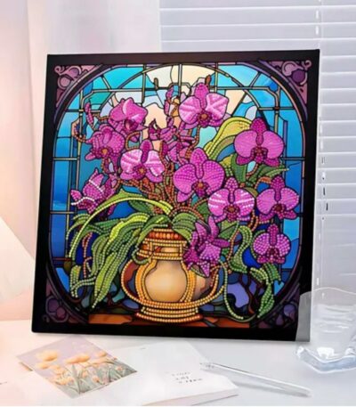 Un tableau de broderie diamant carré, inséré dans un cadre noir, est exposé sur un bureau éclairé. L'œuvre représente un vase doré contenant un grand bouquet d'orchidées roses et violettes avec des feuilles vertes. L'arrière-plan imite un vitrail d'église aux nuances bleu ciel et turquoise, encadré par des arches décoratives sombres. Au premier plan, un verre d'eau transparent et des carnets ouverts entourent le tableau.