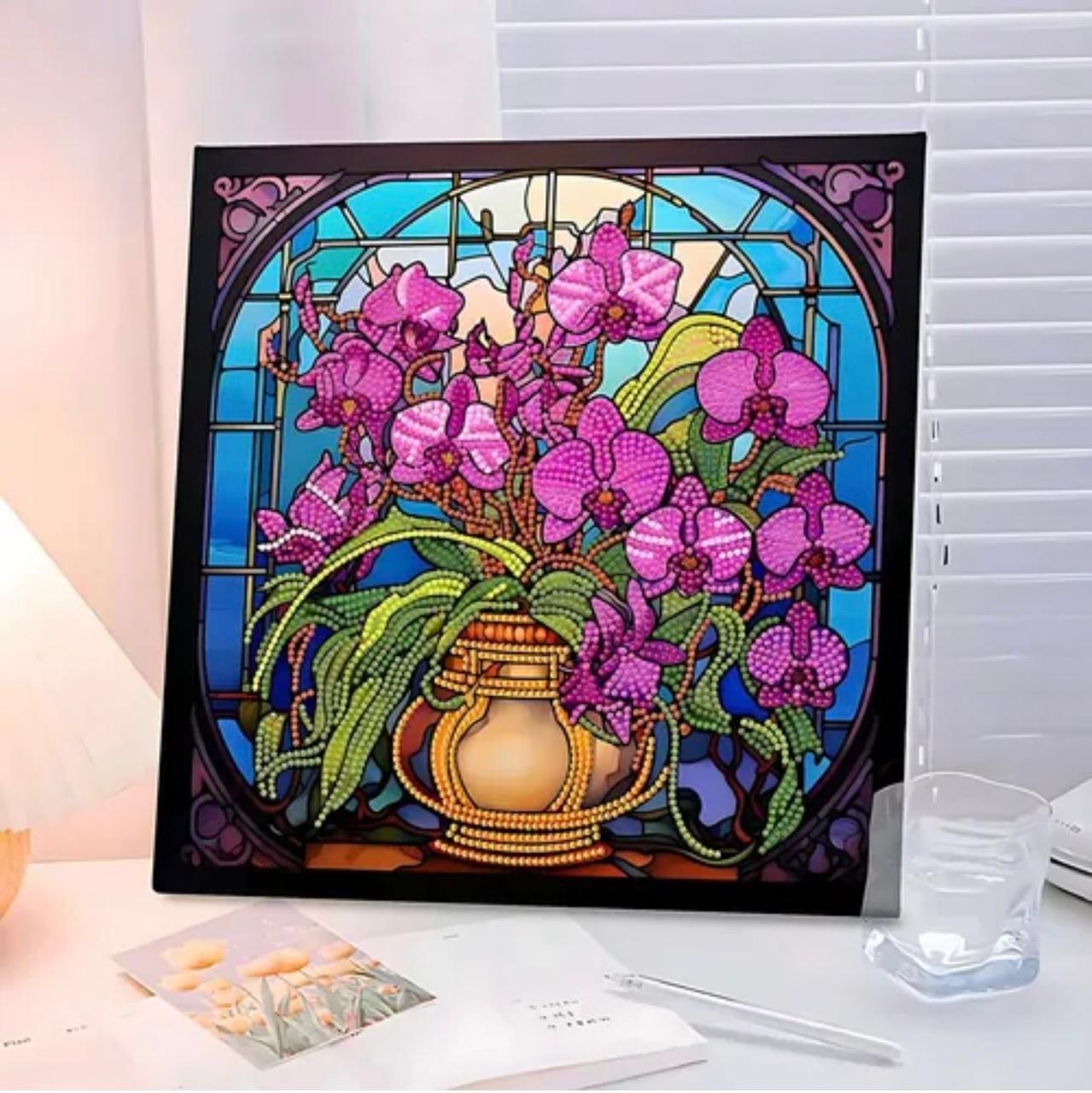 Peinture Diamant Vitrail d'Orchidées Roses en Vase Doré Un tableau de broderie diamant carré, inséré dans un cadre noir, est exposé sur un bureau éclairé. L'œuvre représente un vase doré contenant un grand bouquet d'orchidées roses et violettes avec des feuilles vertes. L'arrière-plan imite un vitrail d'église aux nuances bleu ciel et turquoise, encadré par des arches décoratives sombres. Au premier plan, un verre d'eau transparent et des carnets ouverts entourent le tableau.