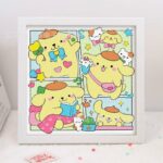 Tableau carré encadré de blanc affichant quatre scènes de style bande dessinée du personnage de Sanrio, Pompompurin. Le personnage, un chien jaune portant son célèbre béret marron, est représenté dans différentes attitudes mignonnes : lisant un livre, tenant un cornet de glace ou saluant joyeusement. L'illustration est entièrement réalisée en broderie diamant, créant une texture scintillante et en relief faite de petits strass colorés.