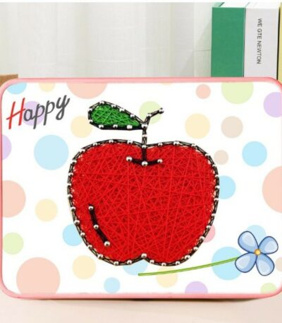 Un tableau en art filaire représentant une grosse pomme rouge sur un support blanc à motifs de gros confettis pastel (bleus, roses, jaunes). La pomme est tissée de fils rouges denses, surmontée d'une petite tige noire et d'une feuille en fils verts. Les contours du fruit sont dessinés par des clous argentés. Le cadre rectangulaire possède des bords arrondis de couleur rose.