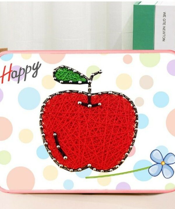 Un tableau en art filaire représentant une grosse pomme rouge sur un support blanc à motifs de gros confettis pastel (bleus, roses, jaunes). La pomme est tissée de fils rouges denses, surmontée d'une petite tige noire et d'une feuille en fils verts. Les contours du fruit sont dessinés par des clous argentés. Le cadre rectangulaire possède des bords arrondis de couleur rose.