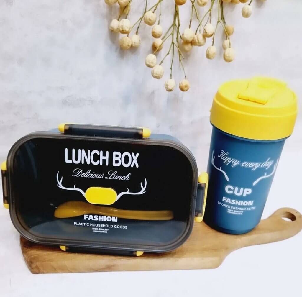 Une lunch box rectangulaire bleu marine avec un couvercle transparent fumé et des clips de fermeture jaunes. Le texte "LUNCH BOX Delicious Lunch" et un logo de bois de cerf blanc ornent le dessus. À côté, un gobelet assorti bleu avec un couvercle jaune arbore l'inscription "CUP Happy every day". L'ensemble repose sur une planche à découper en bois clair devant un mur gris, avec quelques fleurs séchées en arrière-plan.