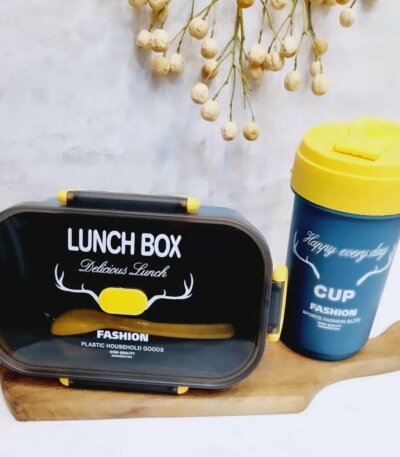 Une lunch box rectangulaire bleu marine avec un couvercle transparent fumé et des clips de fermeture jaunes. Le texte "LUNCH BOX Delicious Lunch" et un logo de bois de cerf blanc ornent le dessus. À côté, un gobelet assorti bleu avec un couvercle jaune arbore l'inscription "CUP Happy every day". L'ensemble repose sur une planche à découper en bois clair devant un mur gris, avec quelques fleurs séchées en arrière-plan.