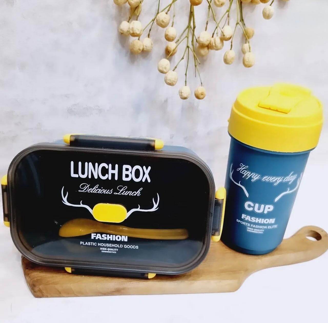 Ensemble Lunch Box "Delicious Lunch" et Gobelet Assorti "Fashion Cup" Une lunch box rectangulaire bleu marine avec un couvercle transparent fumé et des clips de fermeture jaunes. Le texte "LUNCH BOX Delicious Lunch" et un logo de bois de cerf blanc ornent le dessus. À côté, un gobelet assorti bleu avec un couvercle jaune arbore l'inscription "CUP Happy every day". L'ensemble repose sur une planche à découper en bois clair devant un mur gris, avec quelques fleurs séchées en arrière-plan.