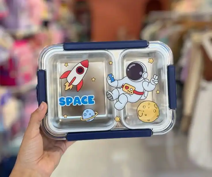 Une boîte à lunch rectangulaire à deux compartiments avec un couvercle transparent orné de motifs spatiaux : un astronaute, une fusée rouge et blanche, une lune jaune et des petites étoiles