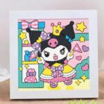 Un kit de broderie diamant terminé et encadré dans un cadre blanc carré posé sur une surface texturée. La toile montre le personnage Kuromi de Sanrio habillée en princesse. Elle porte sa coiffe noire à tête de mort rose surmontée d'une petite couronne jaune, une robe de bal jaune et rose avec un nœud papillon violet à pois blancs, et des chaussures violettes. Kuromi sourit, lève les bras et tient une baguette magique rose. À gauche, une étagère turquoise contient un nœud papillon rose, un flacon bleu et un parfum rose. L'arrière-plan est rose clair à pois blancs, décoré d'étoiles jaunes et de cœurs roses. Toute l'illustration est entièrement recouverte de strass ronds en relief qui donnent un effet texturé et brillant.