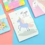 Autour de ce carnet central, on aperçoit les coins d'autres journaux intimes : un modèle jaune vif en haut à gauche avec l'inscription « Just For You », un modèle rose en haut à droite orné d'un grand profil de licorne, et un modèle bleu ciel sur la gauche illustré de motifs floraux.