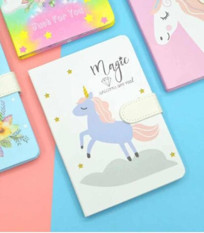 Autour de ce carnet central, on aperçoit les coins d'autres journaux intimes : un modèle jaune vif en haut à gauche avec l'inscription « Just For You », un modèle rose en haut à droite orné d'un grand profil de licorne, et un modèle bleu ciel sur la gauche illustré de motifs floraux.