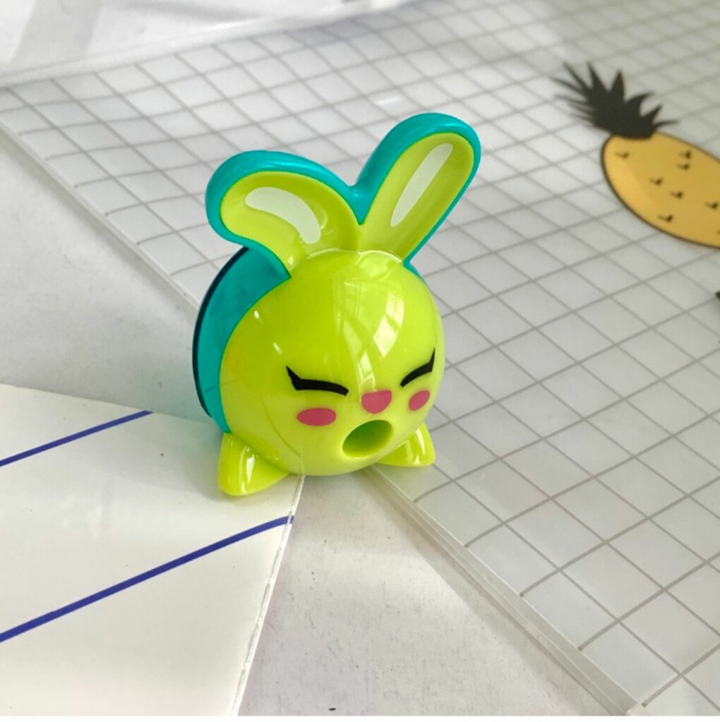 Un taille-crayon en plastique de la marque Maped en forme de tête de lapin rigolo, de couleurs vert pomme et bleu turquoise. Les oreilles du lapin servent de poignées et l'ouverture pour insérer le crayon se situe au niveau de sa bouche grande ouverte. Le taille-crayon est posé sur un cahier à petits carreaux.