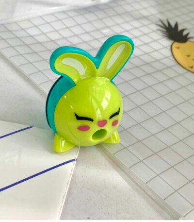 Un taille-crayon en plastique de la marque Maped en forme de tête de lapin rigolo, de couleurs vert pomme et bleu turquoise. Les oreilles du lapin servent de poignées et l'ouverture pour insérer le crayon se situe au niveau de sa bouche grande ouverte. Le taille-crayon est posé sur un cahier à petits carreaux.