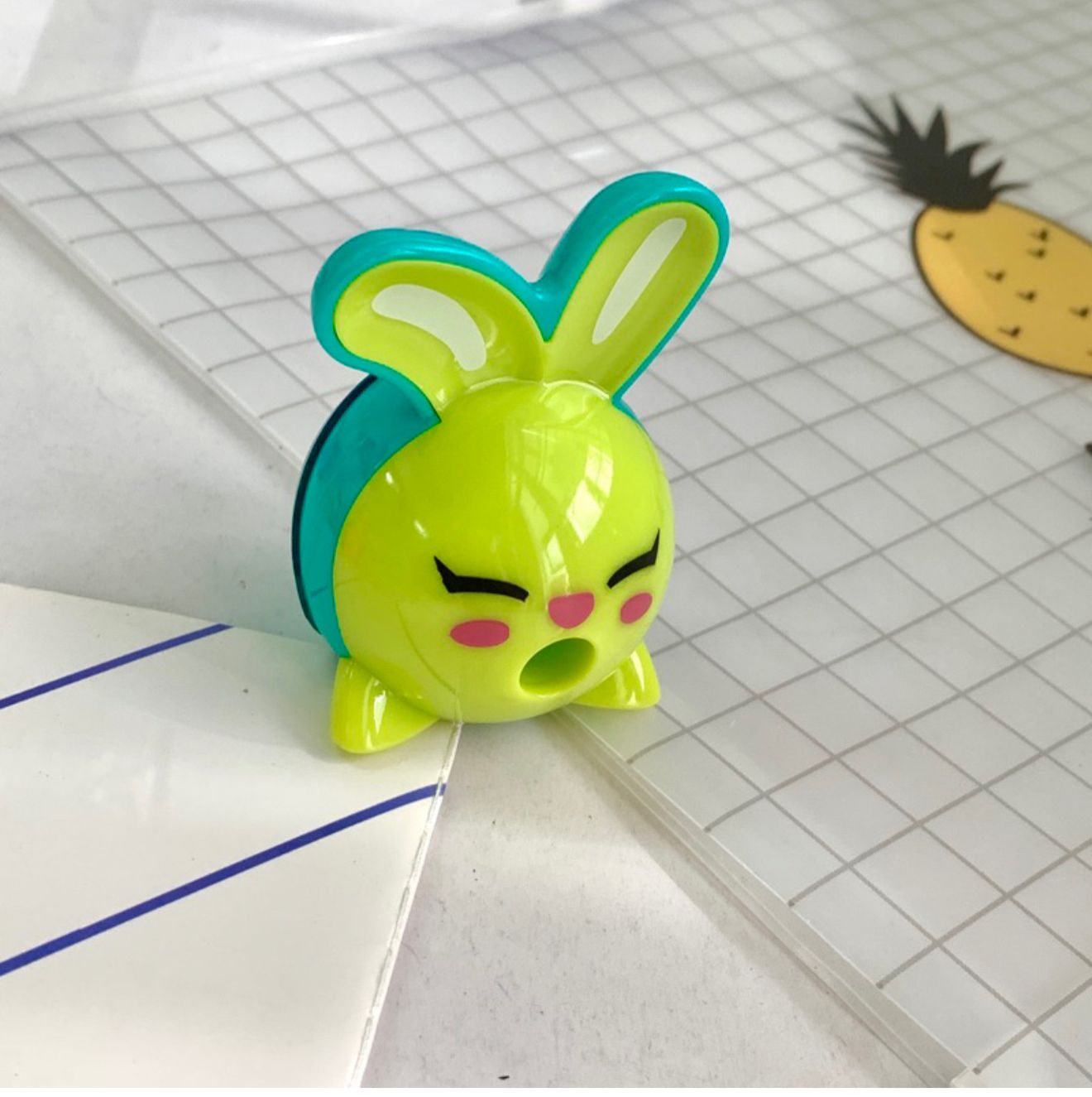 Taille-crayons Lapin Maped Croc Croc Innovation Vert/Turquoise. Un taille-crayon en plastique de la marque Maped en forme de tête de lapin rigolo, de couleurs vert pomme et bleu turquoise. Les oreilles du lapin servent de poignées et l'ouverture pour insérer le crayon se situe au niveau de sa bouche grande ouverte. Le taille-crayon est posé sur un cahier à petits carreaux.