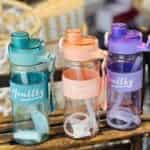 Trois gourdes de sport transparentes de la marque Healthy, déclinées en bleu turquoise, orange pêche et violet. Elles disposent d'un manchon en silicone strié assorti, d'une sangle de transport et d'une paille blanche visible à l'intérieur.