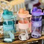 Trois gourdes de sport transparentes de la marque Healthy, déclinées en bleu turquoise, orange pêche et violet. Elles disposent d'un manchon en silicone strié assorti, d'une sangle de transport et d'une paille blanche visible à l'intérieur.