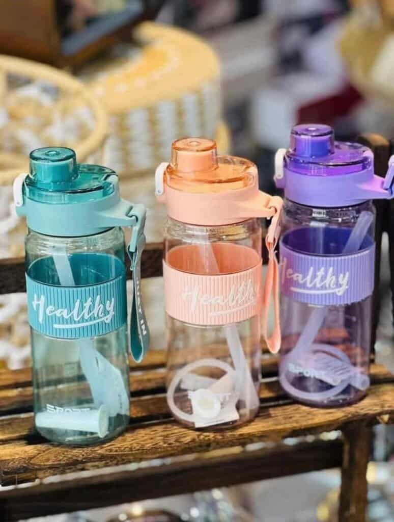 Trois gourdes de sport transparentes de la marque Healthy, déclinées en bleu turquoise, orange pêche et violet. Elles disposent d'un manchon en silicone strié assorti, d'une sangle de transport et d'une paille blanche visible à l'intérieur.