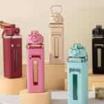 Cinq bouteilles d'eau de forme carrée disposées verticalement sur des socles cylindriques. Chaque bouteille possède un corps en plastique transparent coloré (rouge brique, rose, crème, bleu ciel et noir) partiellement recouvert d'un manchon de protection texturé assorti. Les manchons présentent un motif en relief et une fenêtre verticale transparente permettant de voir le niveau de liquide.