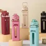Cinq bouteilles d'eau de forme carrée disposées verticalement sur des socles cylindriques. Chaque bouteille possède un corps en plastique transparent coloré (rouge brique, rose, crème, bleu ciel et noir) partiellement recouvert d'un manchon de protection texturé assorti. Les manchons présentent un motif en relief et une fenêtre verticale transparente permettant de voir le niveau de liquide.